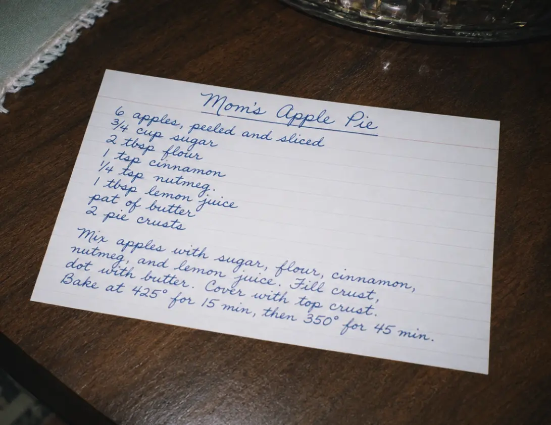 A handwritten apple pie recipe card on a worn wooden kitchen counter, warm afternoon light streaming in from a nearby window.